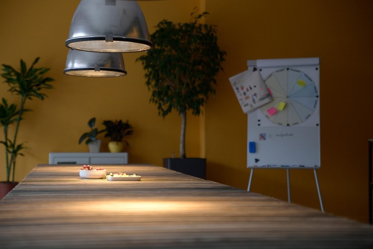 Table de réunion éclairée par lampe suspendue, paperboard avec schémas et sticky notes en arrière-plan, plantes vertes Table de réunion éclairée par lampe suspendue, paperboard avec schémas et sticky notes en arrière-plan, plantes vertes